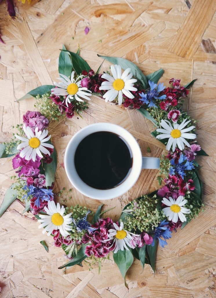 A cup of black coffee on a wooden table encircled by a wreath of colorful flowers.