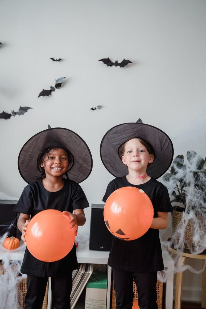 Two kids in witch costumes enjoying Halloween indoors with decorations and balloons.